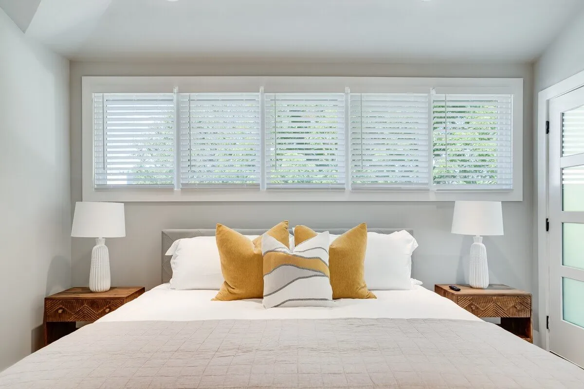 A serene bedroom with a cozy bed adorned with decorative pillows, lamps on matching nightstands, and a window fitted with blinds to provide privacy and darkness for restful sleep.