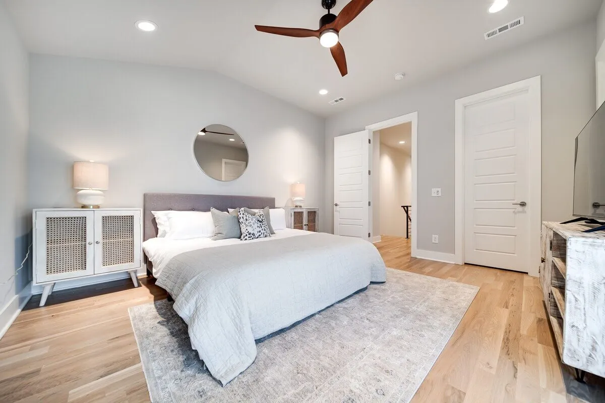A tranquil bedroom featuring a bed with soft gray bedding, a round mirror above a wooden nightstand with a lamp, wooden flooring, and a decorative rug. Natural light floods the space, creating a soothing environment ideal for relaxation or quiet moments.