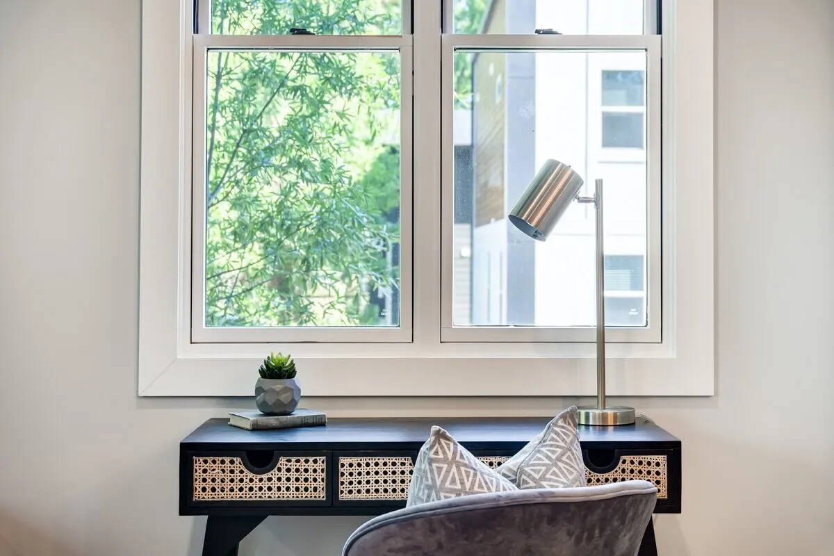 A calm, sunlit workspace featuring a desk and lamp by the window, surrounded by greenery and soft pillows. The natural light streams in, creating an inviting and productive atmosphere ideal for both work and relaxation.