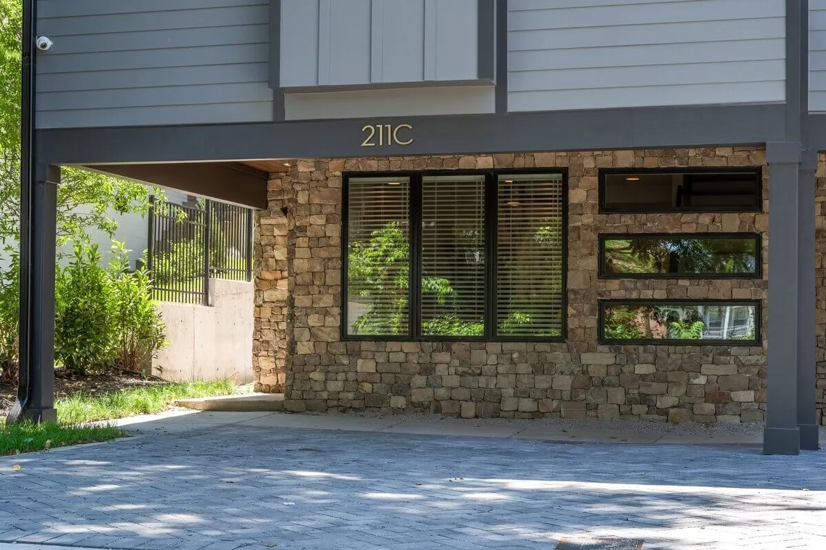 The image shows the exterior of a contemporary home with a stone wall, large windows, and the house number '211C' clearly displayed. There is a sense of arrival, with a well-kept landscape and a welcoming front entrance.