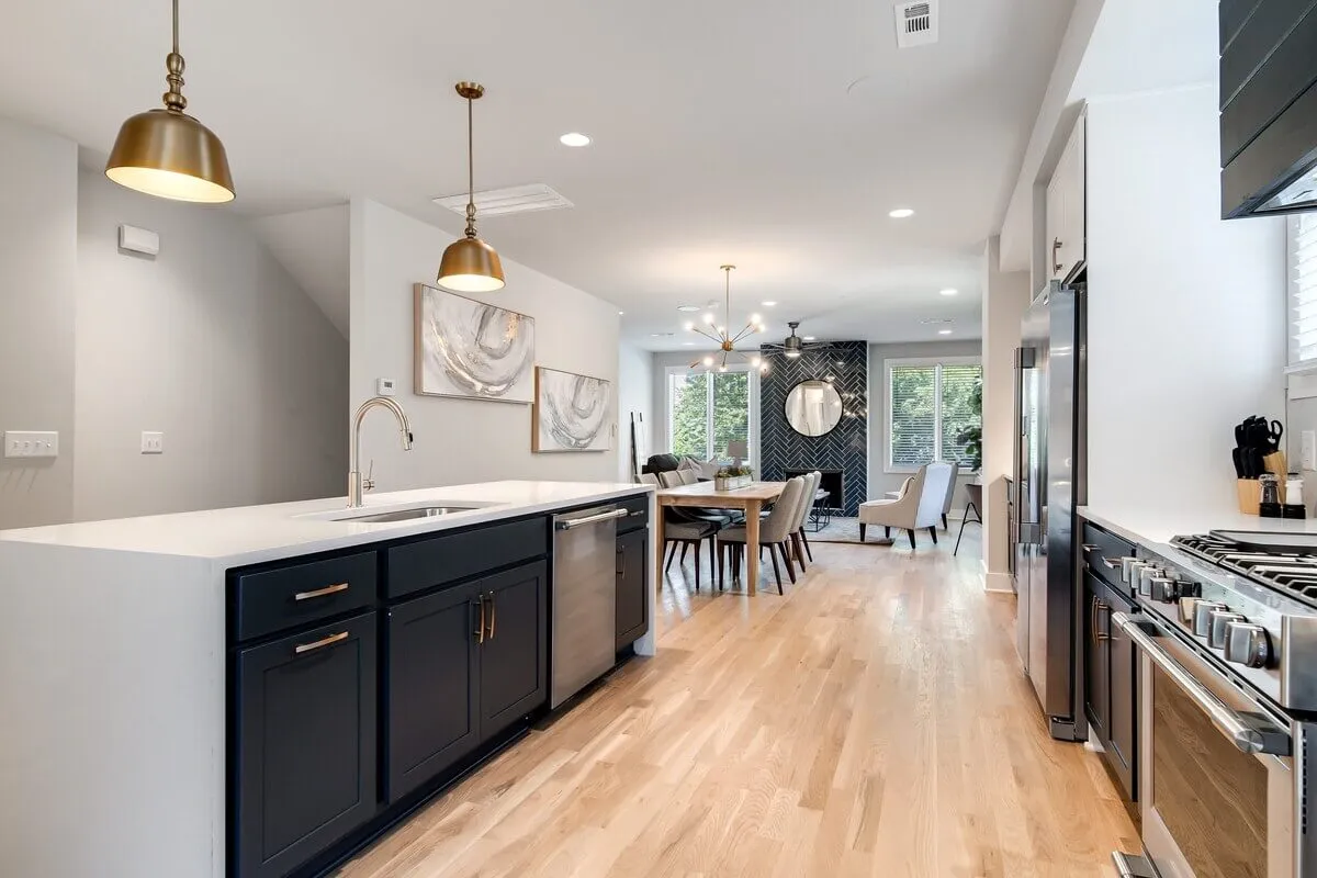 A stylish interior shot capturing a spacious kitchen with a central island flowing seamlessly into the dining area. Pendant lights illuminate the workspace, and modern artwork adds a vibrant touch. The scene hints at both cooking and socializing possibilities, with sleek surfaces and ample prep space visible.