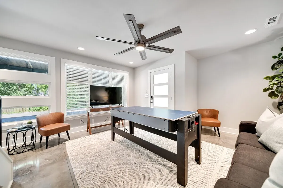 A spacious, light-filled game room featuring a ping pong table, sofa, television, and large windows with views outside. The space exudes a playful, casual vibe perfect for family bonding or unwinding after a busy day.