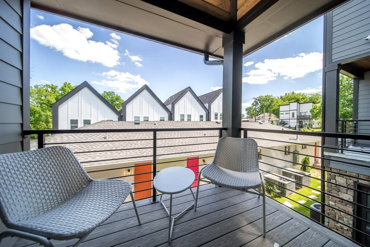 Two comfortable outdoor chairs and a small round table are arranged on a balcony or patio, with a view overlooking neighboring buildings. The sun-soaked setting invites quiet moments, morning coffee, or evening conversations and highlights the urban context while maintaining a sense of refuge.