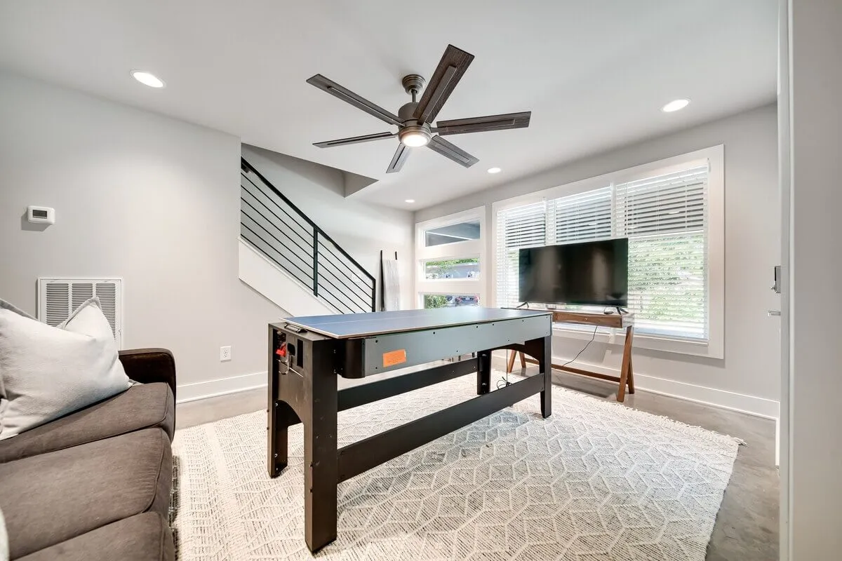 The image shows a spacious entertainment area equipped with an air hockey table, sofa, television, ceiling fan, and a staircase leading to other parts of the home. It's a versatile space for both lively gatherings and relaxing after an eventful day.