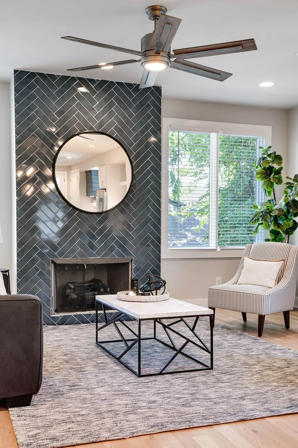 A cozy, modern lounge area featuring a chevron tiled wall, circular mirror, elegant fireplace, coffee table, and comfortable striped armchair. The scene is warm and welcoming, ideal for intimate conversations and casual gatherings.