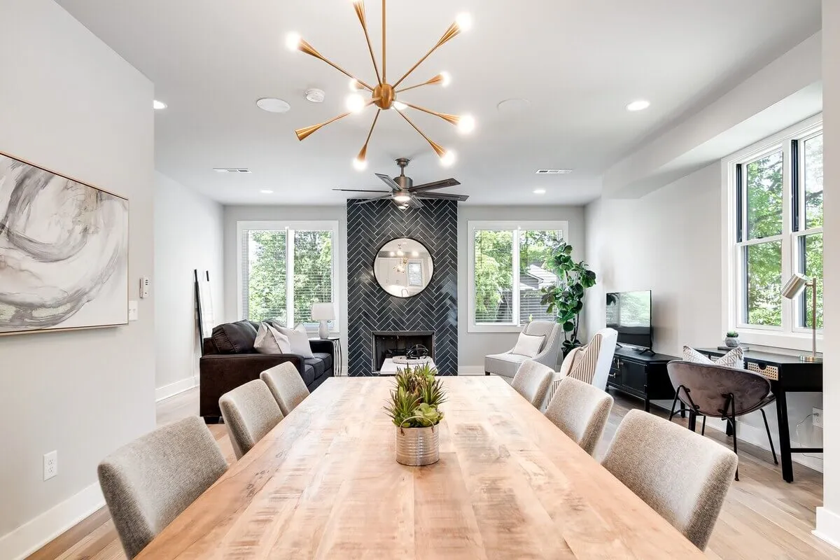 The image showcases an elegant, sun-filled dining area with a sophisticated chandelier overhead, a modern fireplace, and large windows that fill the space with natural light. The inviting seating area promises comfort, making it perfect for shared meals, celebrations, or relaxation.