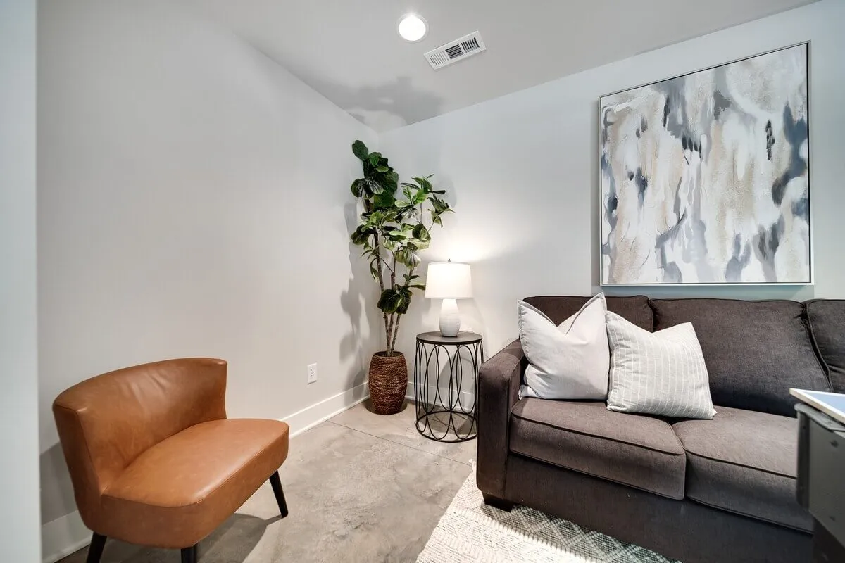 A comfortable seating area with a plush sofa, accent chair, floor lamp, lush plant, and wall art. The space feels welcoming and is ideal for relaxing or having informal conversations after a workday.